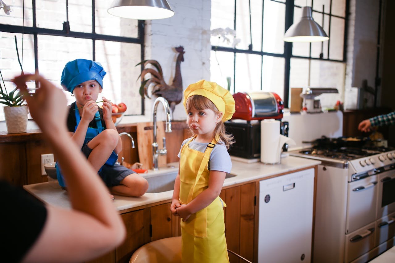 Two kids in colorful chef hats enjoy a playful cooking session in a cozy kitchen setting.