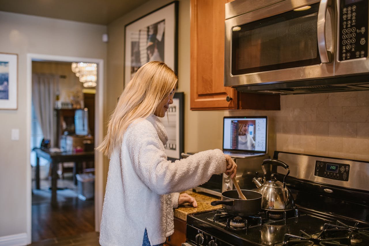 A woman cooks while attending an online class from her laptop in the cozy kitchen.