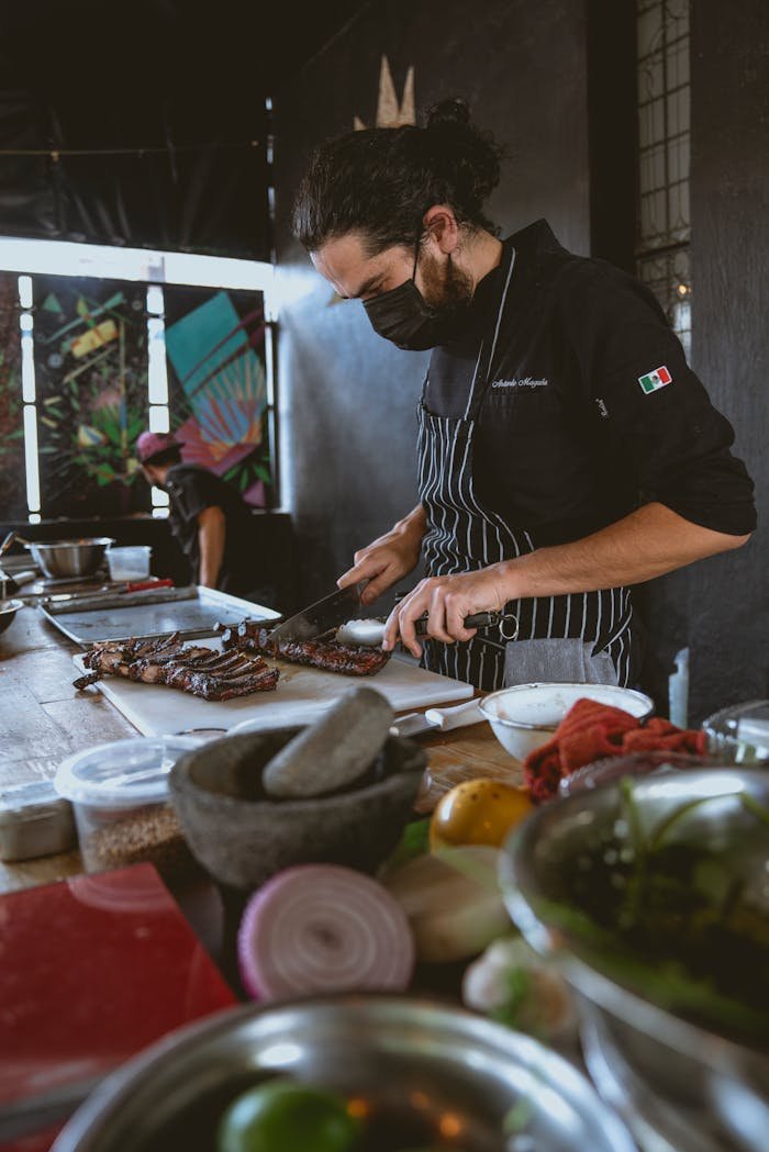 Masked chef prepares steak in a vibrant indoor culinary workshop, showcasing professional cooking skills.