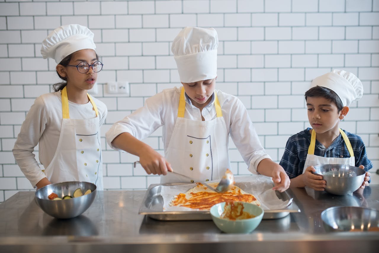 Three children in chef hats and aprons preparing a meal in an indoor kitchen setting.
